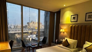 Ein Hotelzimmer mit zwei Betten, einem Schreibtisch und einem Sessel. Ein großes Fenster bietet Blick auf die Stadt.