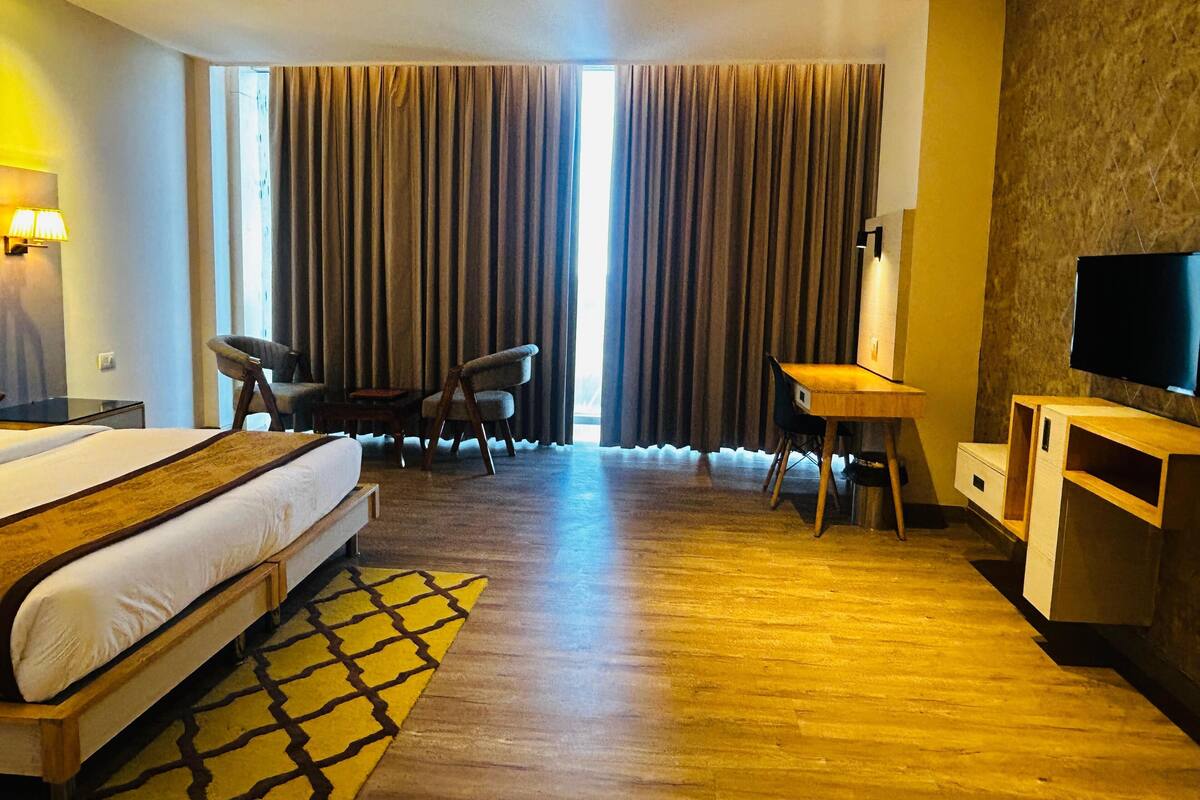Une chambre d’hôtel comprenant un lit, deux chaises, un bureau, une télévision et une grande fenêtre avec des rideaux.