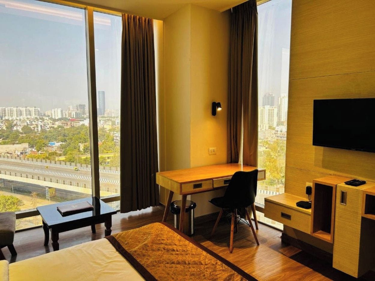 A hotel room with a large window, a desk, a chair, a bed, and a TV.