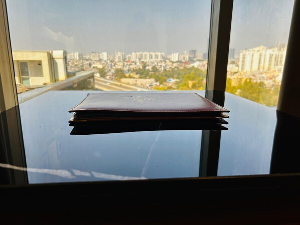 A book on a glass table by a window with a city view. - Hotel Sakura Gold (Gurugram)