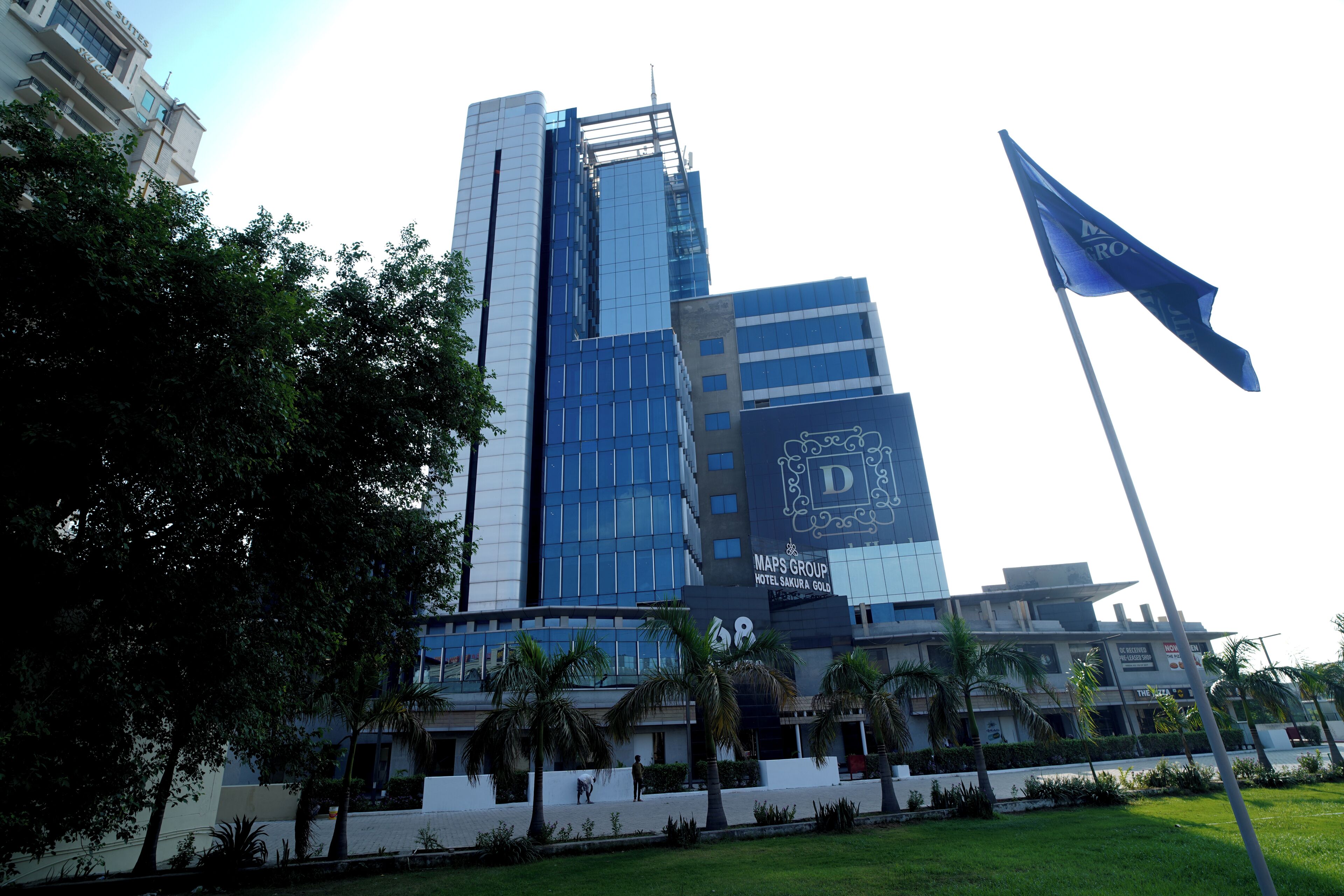 A modern hotel with a prominent glass facade, a flagpole with a blue flag, and a well-maintained lawn with palm trees.