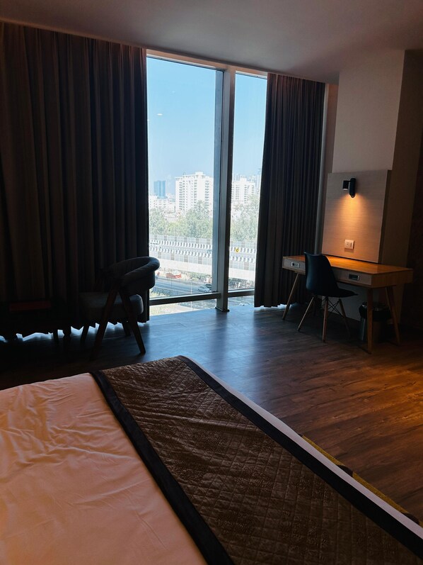 A hotel room with a bed, a desk, and a view of the city through large windows. - Hotel Sakura Gold (Gurugram)
