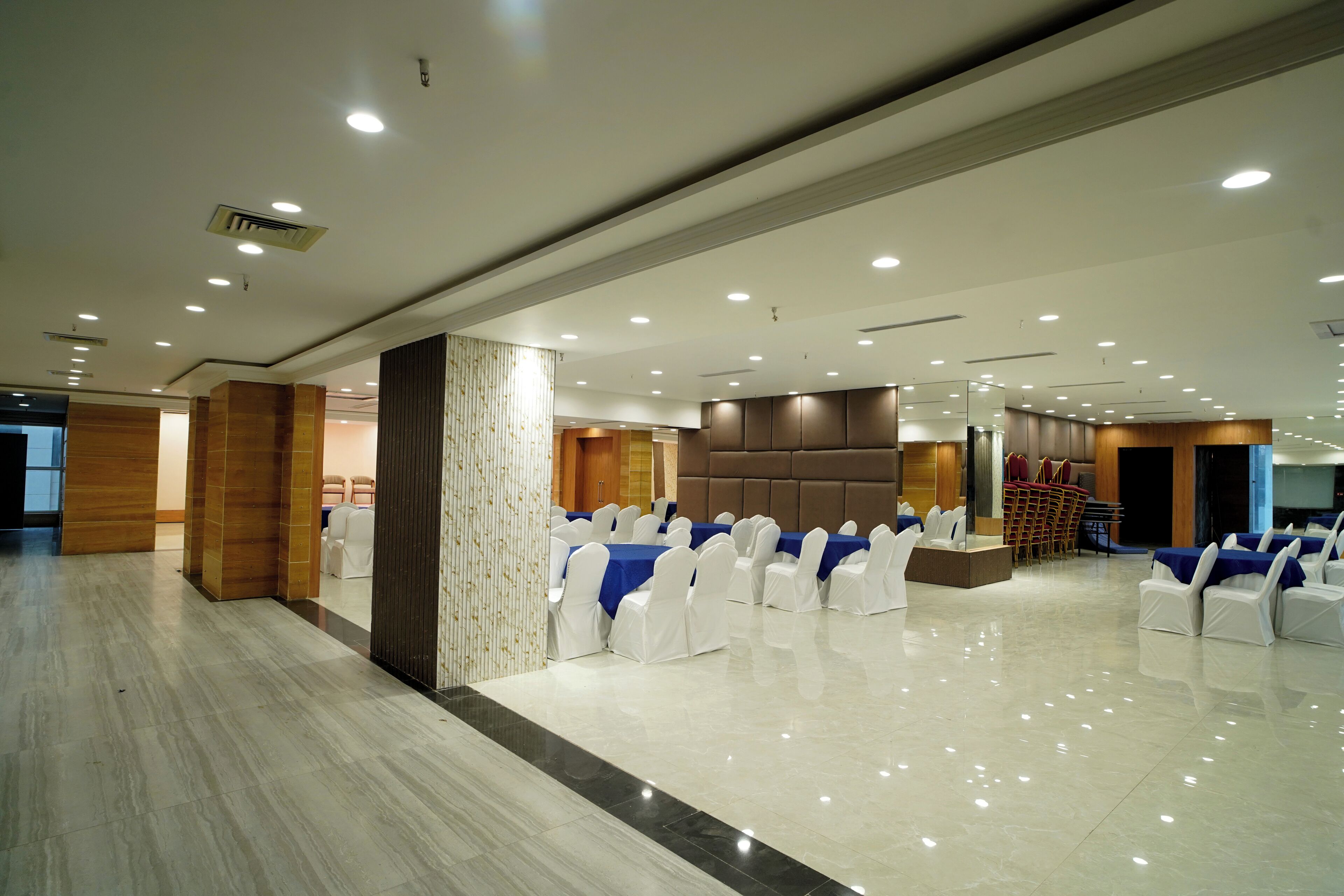 A spacious banquet hall with white chairs and blue tablecloths, a polished marble floor, and recessed ceiling lighting.