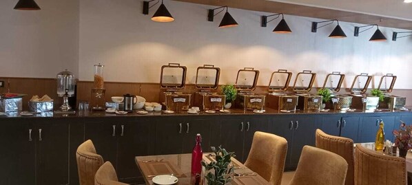 A breakfast bar with multiple serving stations, a dining area with chairs and a table, and a wall-mounted light fixture. - Hotel Sakura Gold (Gurugram)