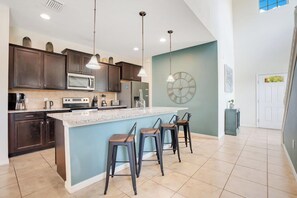 A modern kitchen with dark cabinets, a large island, bar stools, pendant lighting, and a clock on the wall.