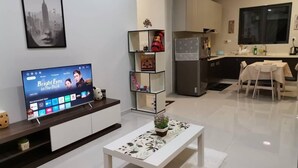 A modern living room with a flat-screen TV displaying a movie, a white coffee table with a plant, and a kitchen area with a refrigerator and dining table. - SeaView Country Garden 3BR2FREE By Natol (Johor Bahru)