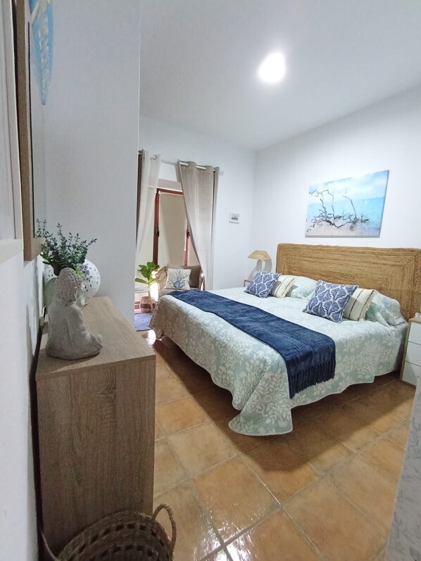 Een slaapkamer met een bed, nachtkastje, lamp, wanddecoratie en een beeldje op een houten dressoir.