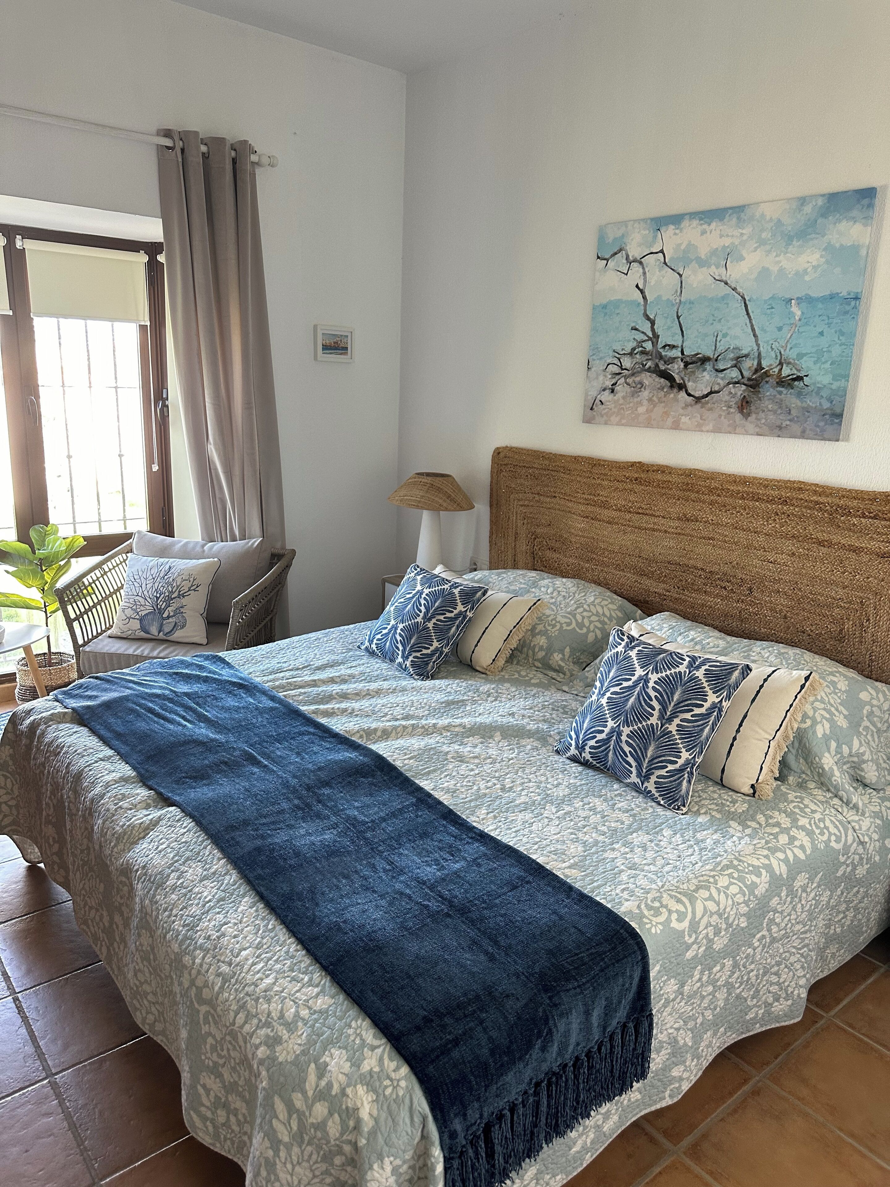 A bedroom with a large bed, a bedside table with a lamp, a window with curtains, and a painting on the wall.