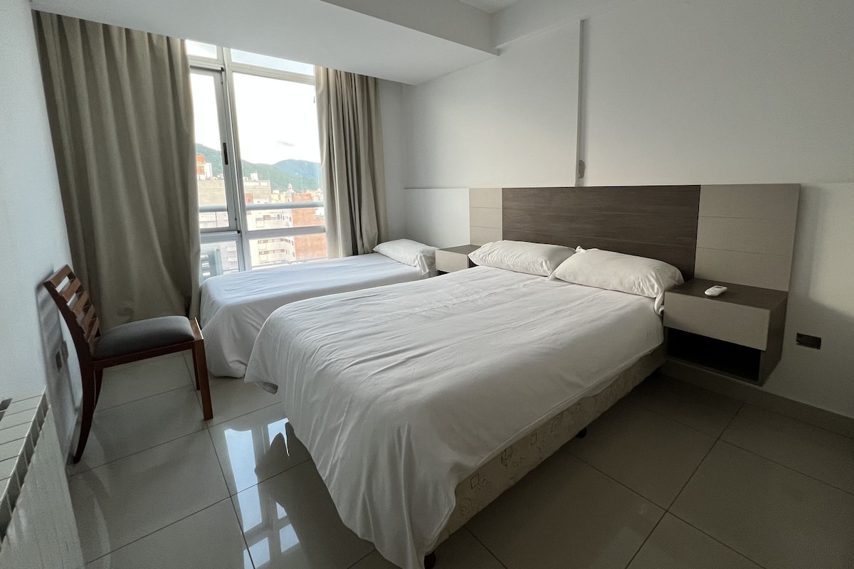 Habitación de hotel con dos camas, una silla, una mesita de noche y vistas a la ciudad a través de amplias ventanas.