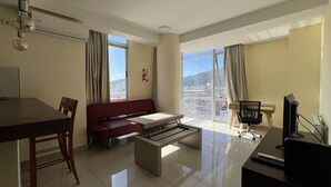 Una sala de estar moderna con sofá, mesa de comedor y televisor. Amplias ventanas ofrecen vista a la ciudad.