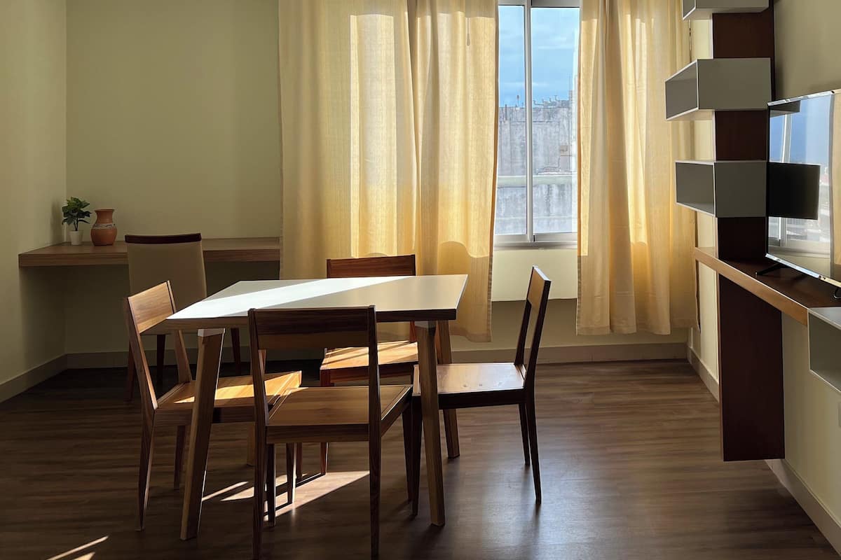 Un comedor con una mesa y sillas de madera, una planta en maceta y vistas a través de una ventana con cortinas.
