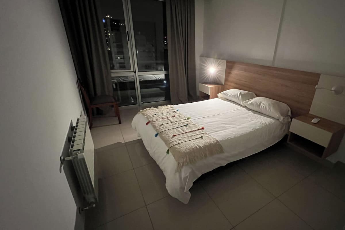 Habitación de hotel con una cama grande, una mesita de noche, una lámpara, una silla y vista a la ciudad por la ventana.