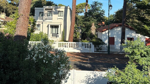 A two-story building with a white facade, surrounded by tall pine trees and a white picket fence.