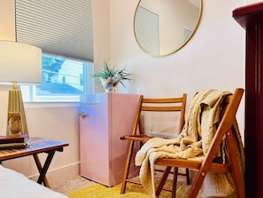 A room with a pink refrigerator, a wooden chair with a blanket, a small table with a lamp, and a window with blinds.