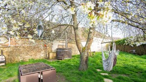 Un jardin avec un mur en pierre, un barbecue, un tonneau en bois, une chaise suspendue et un hamac.