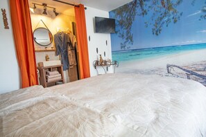 Une chambre avec un lit, une salle de bain avec un lavabo et un miroir, et une fresque représentant une plage sur le mur.