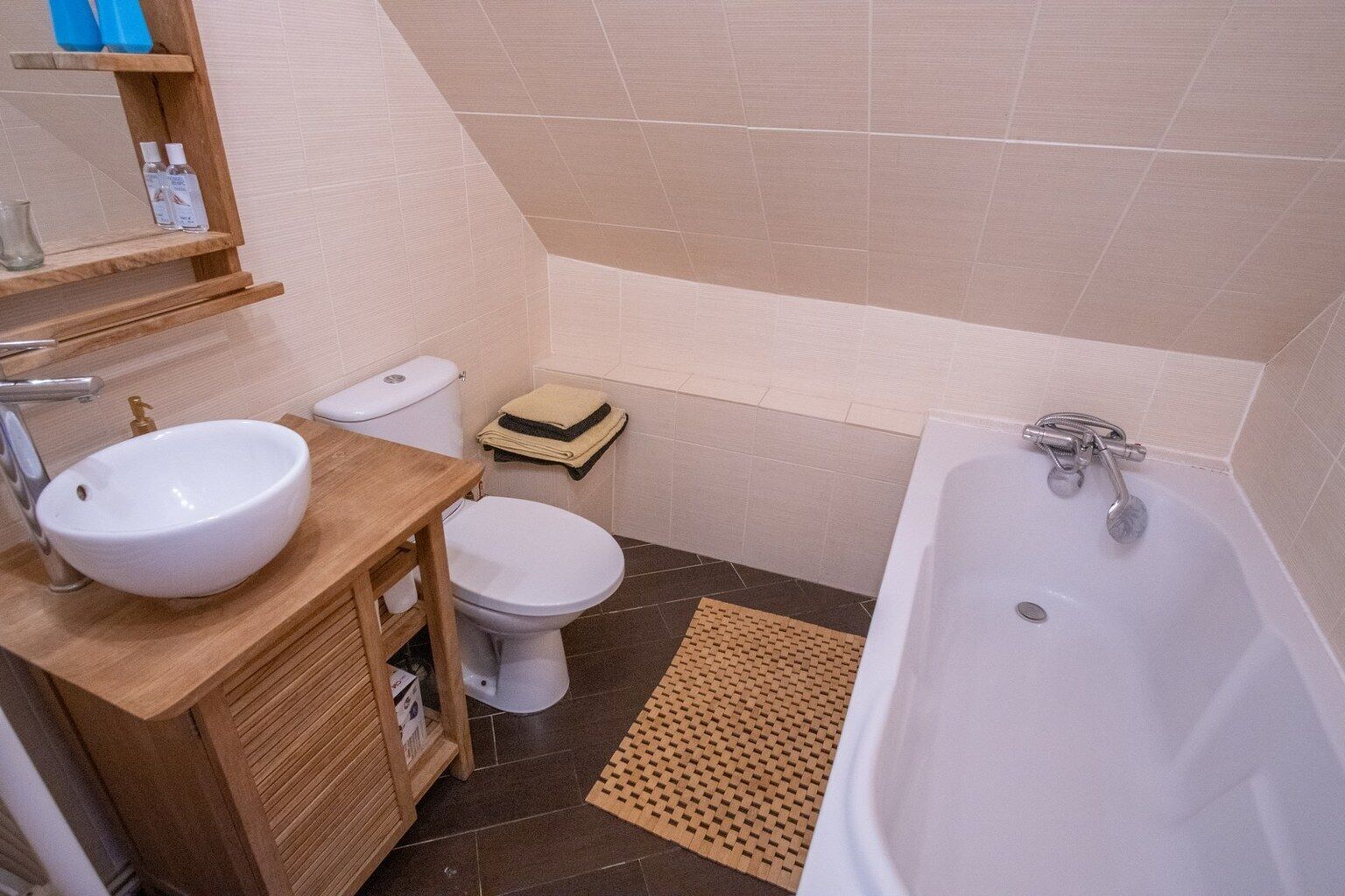 A bathroom with a white sink, toilet, and bathtub. There is a wooden vanity with a mirror and a shelf with toiletries.