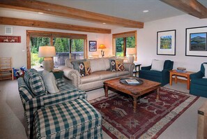 A cozy living room with a sofa, two armchairs, a coffee table, and a rug. There are two windows with curtains, a lamp, and framed pictures on the wall. - Creek Cove, Gorgeous home, hot tub, very private (Crested Butte)
