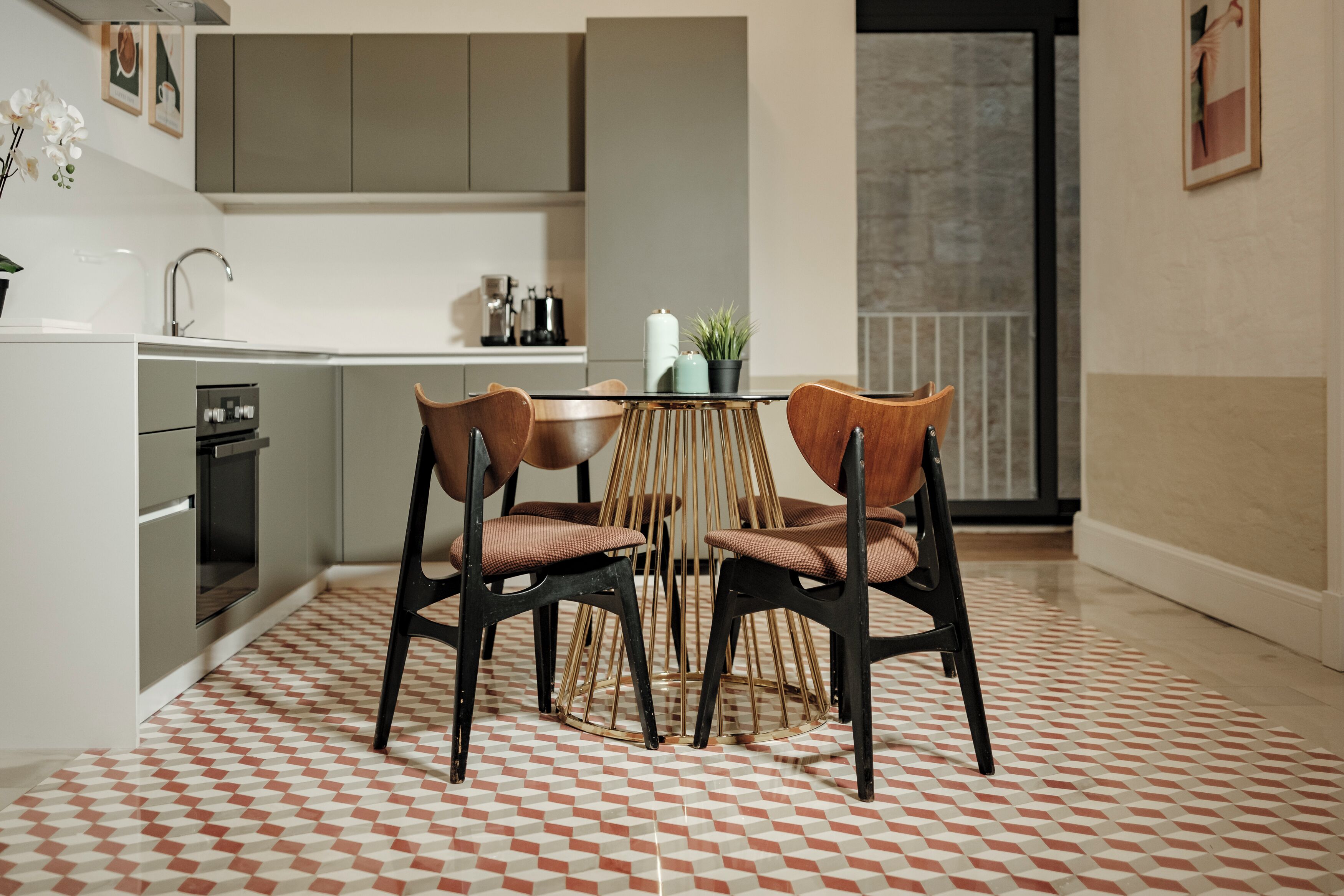A modern kitchen with a dining area. It includes a kitchen counter, a sink, and a stove. There are two wooden chairs with cushions around a small round table. The floor has a geometric patterned tile design.