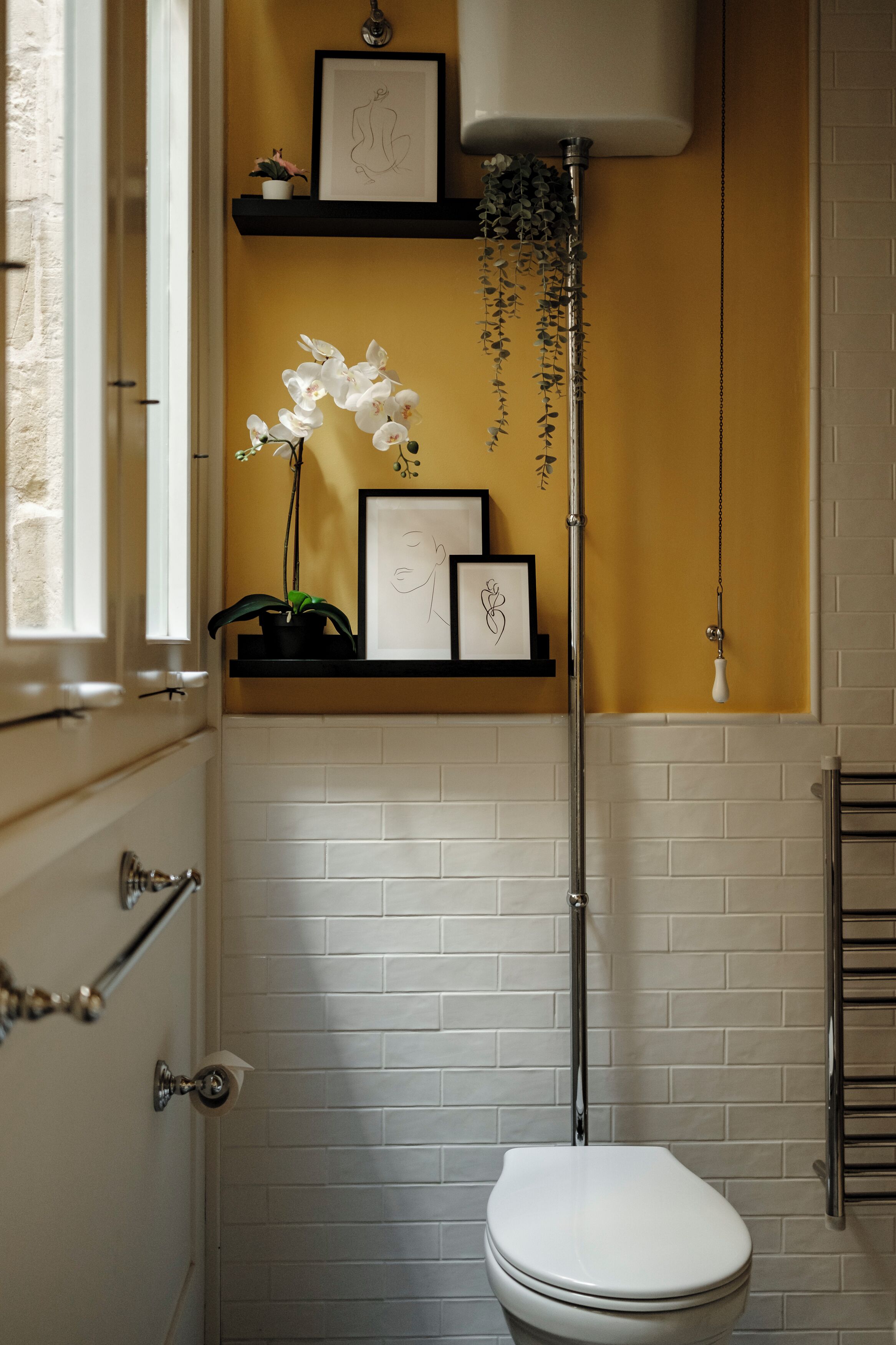 Une salle de bain avec des toilettes blanches, un lavabo avec un robinet chromé, une étagère avec des œuvres d’art encadrées et une plante en pot, ainsi qu’un luminaire suspendu.