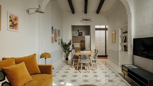 Ein Wohnzimmer mit einer Couch, einem Esstisch mit Stühlen, einem Fernseher und einer Küche im Hintergrund.