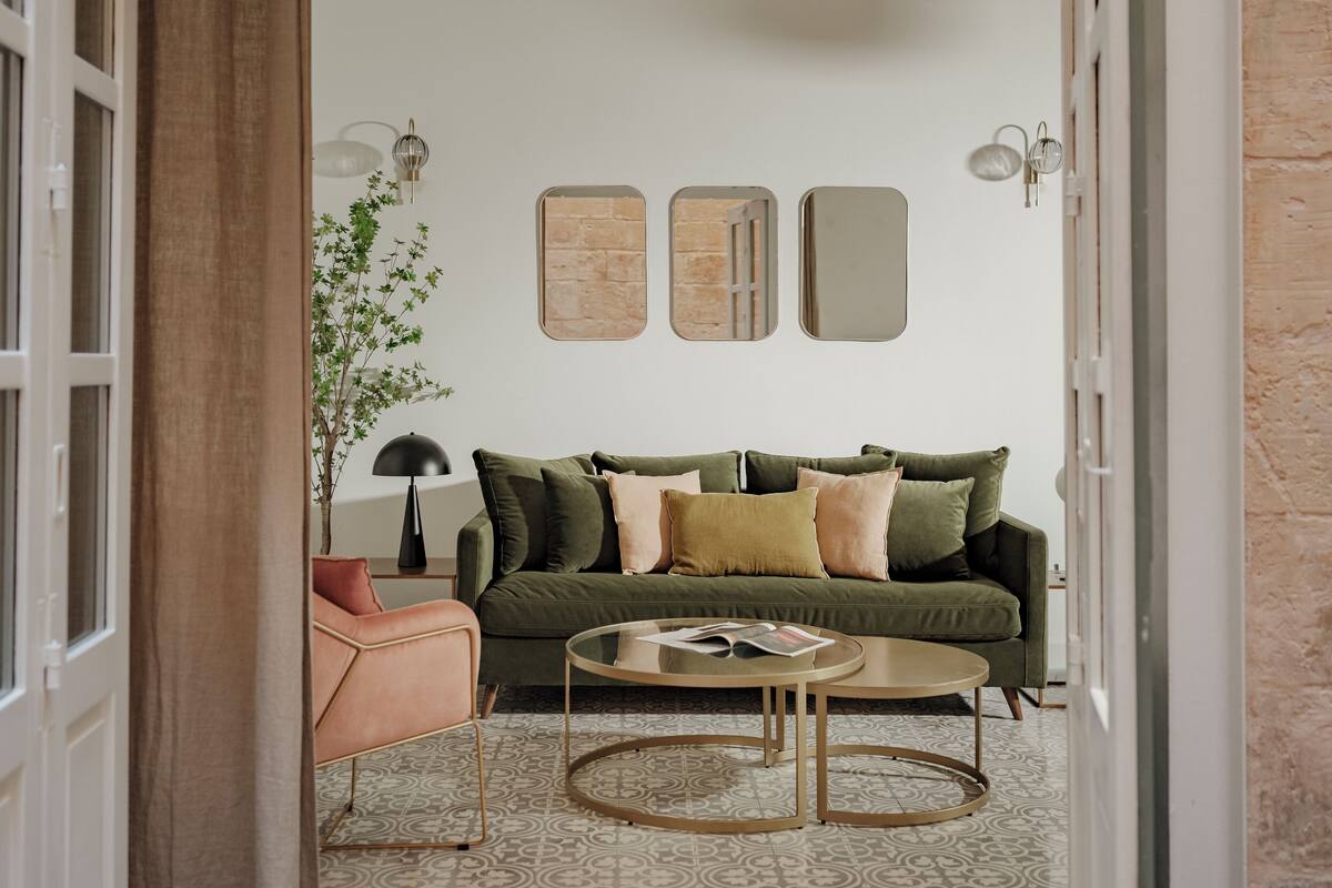 Uma sala de estar com um sofá verde, uma mesa de centro redonda e uma poltrona rosa. Há três espelhos fixados na parede e uma luminária sobre uma mesinha de apoio.