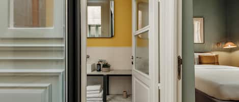 A hotel room with a bed, a bedside table, a mirror, and a bathroom with a sink and towel rack.