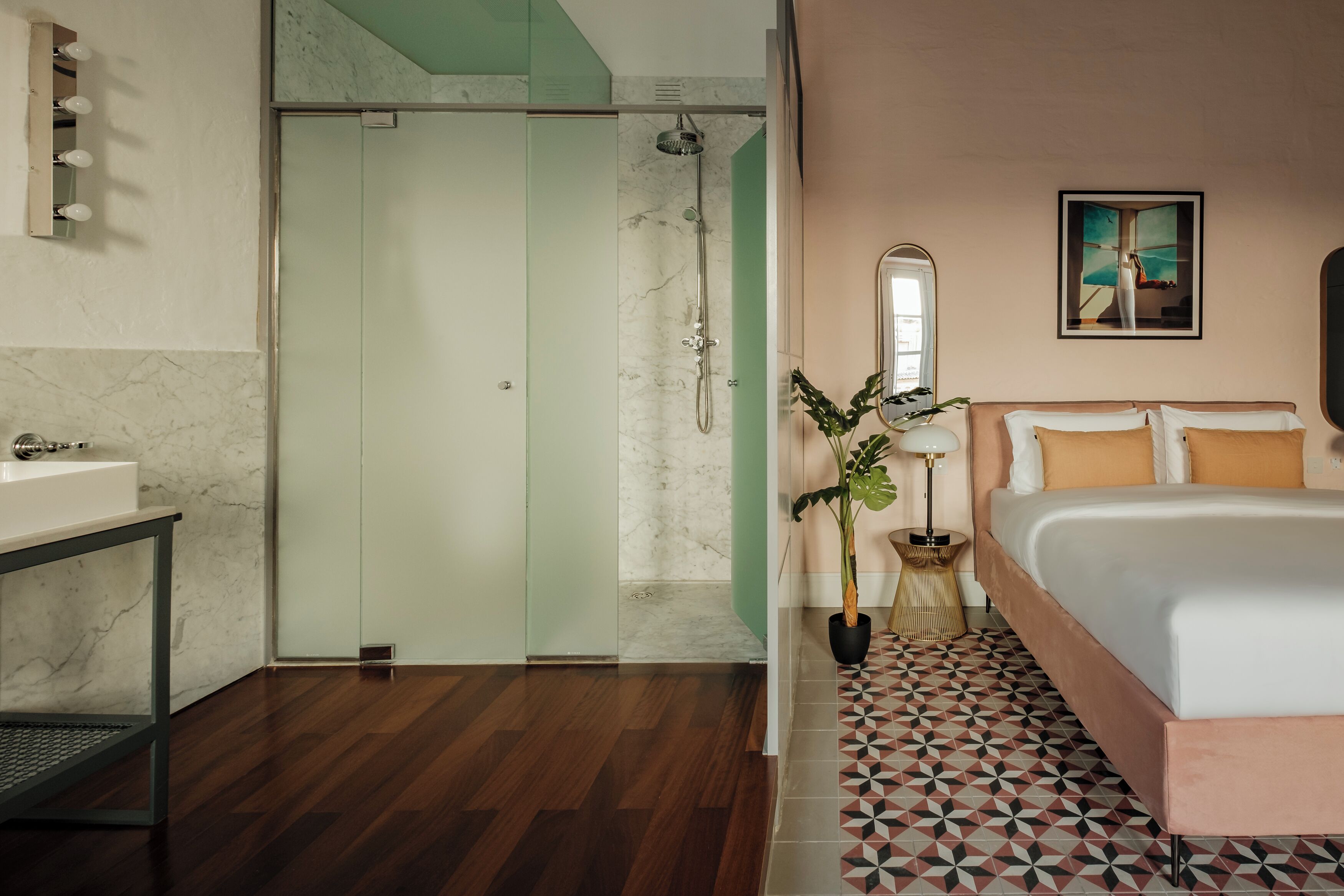 Een moderne hotelkamer met een douchecabine van glas, een bed met vloer in geometrisch patroon, een nachtkastje, een spiegel en een pot met een plant.