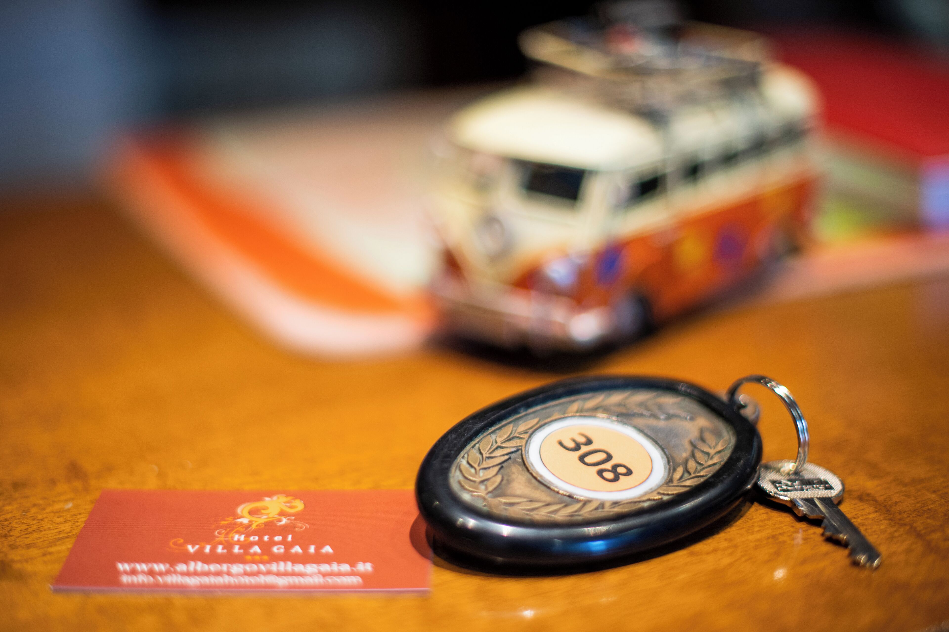 A key fob with the number 308 on a wooden surface, a business card for Hotel Villagaja, and a blurred toy car in the background.