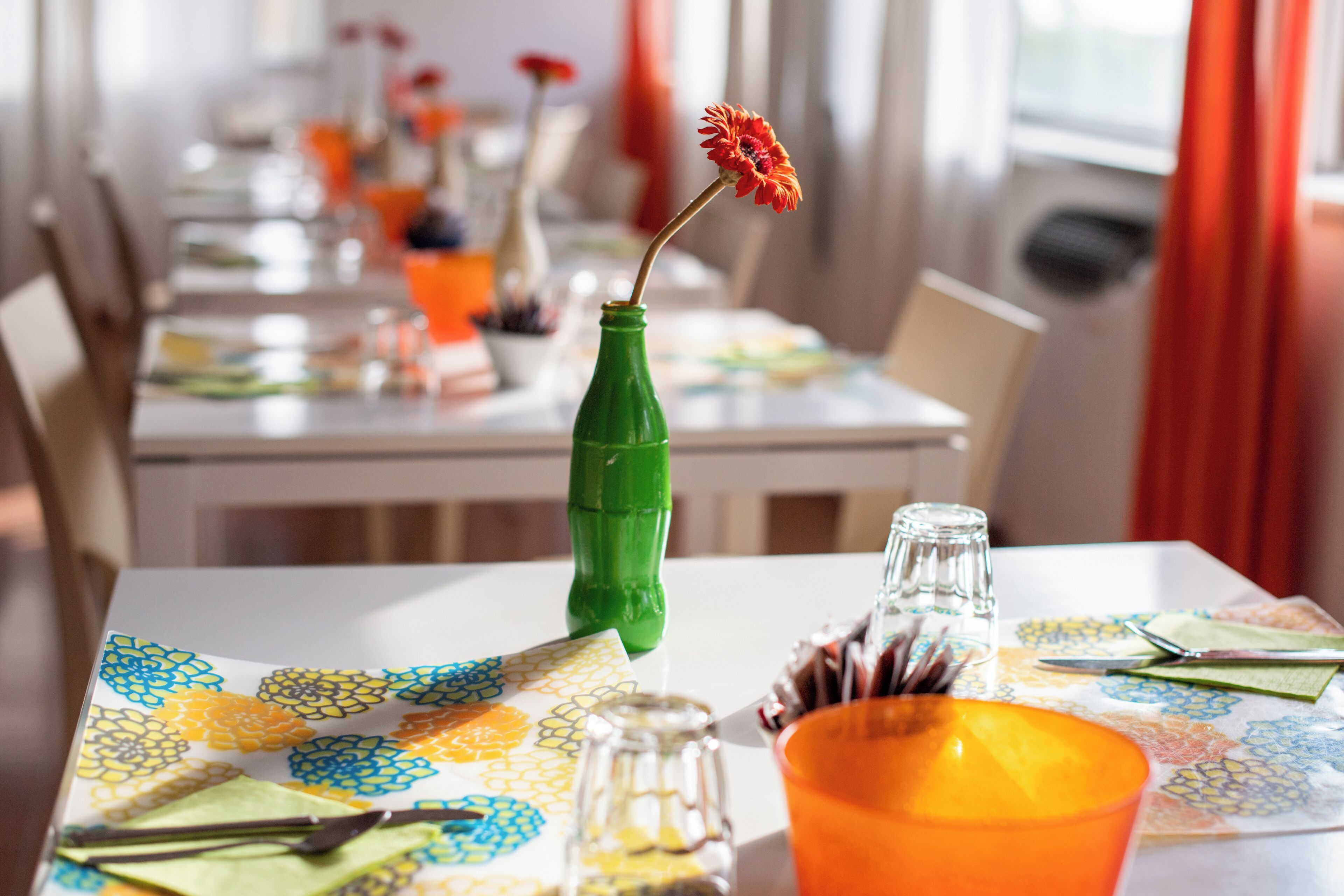 Une table à manger recouverte d’une nappe colorée, une bouteille verte servant de vase et contenant une seule fleur, et un salier en verre.