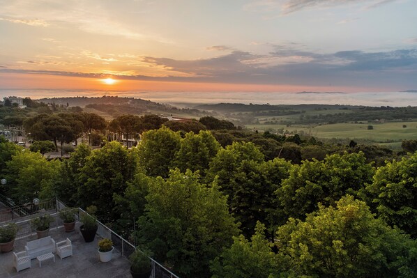 Een terras met buiten zitplaatsen en bloempotten, met uitzicht op een weelderig groonlandschap en een zonsondergang.