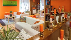 A modern living room with a white sofa, orange cushions, a wooden coffee table, and a bookshelf with decorative items and books. - Albergo Villa Gaia (Chianciano Terme)