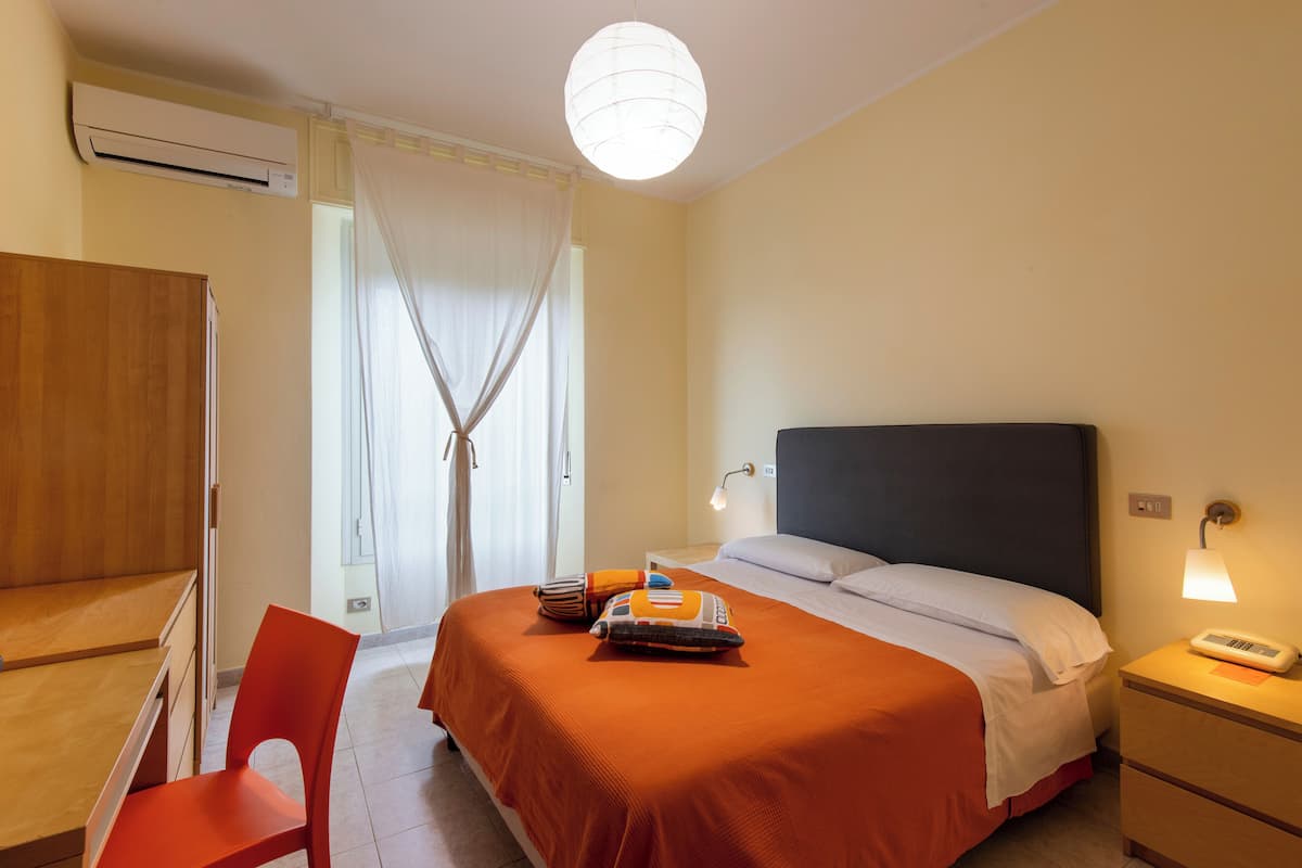 Hotellihuone, jossa on sänky, yöpöytä, lamppu ja työpöytä tuolilla.
