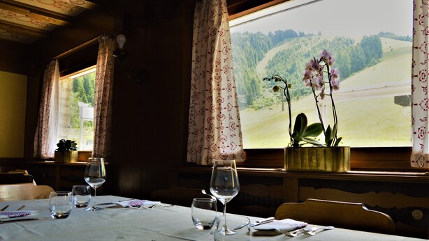 A dining table set with cutlery and wine glasses, a vase with flowers, and a window with a view of a green landscape. - Hotel Cristallo (Folgaria)