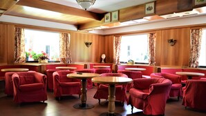 A restaurant with red upholstered chairs and round tables, wooden paneling, and large windows with curtains. - Hotel Cristallo (Folgaria)