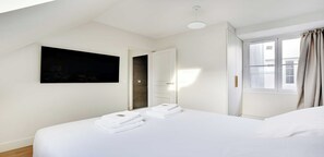 A hotel room with a large bed, a flat-screen TV mounted on the wall, a window with curtains, and a door leading to another room. - Bright & Stylish Two-Bedroom Apartment with Modern Comforts in Saint-Germain (Paris)