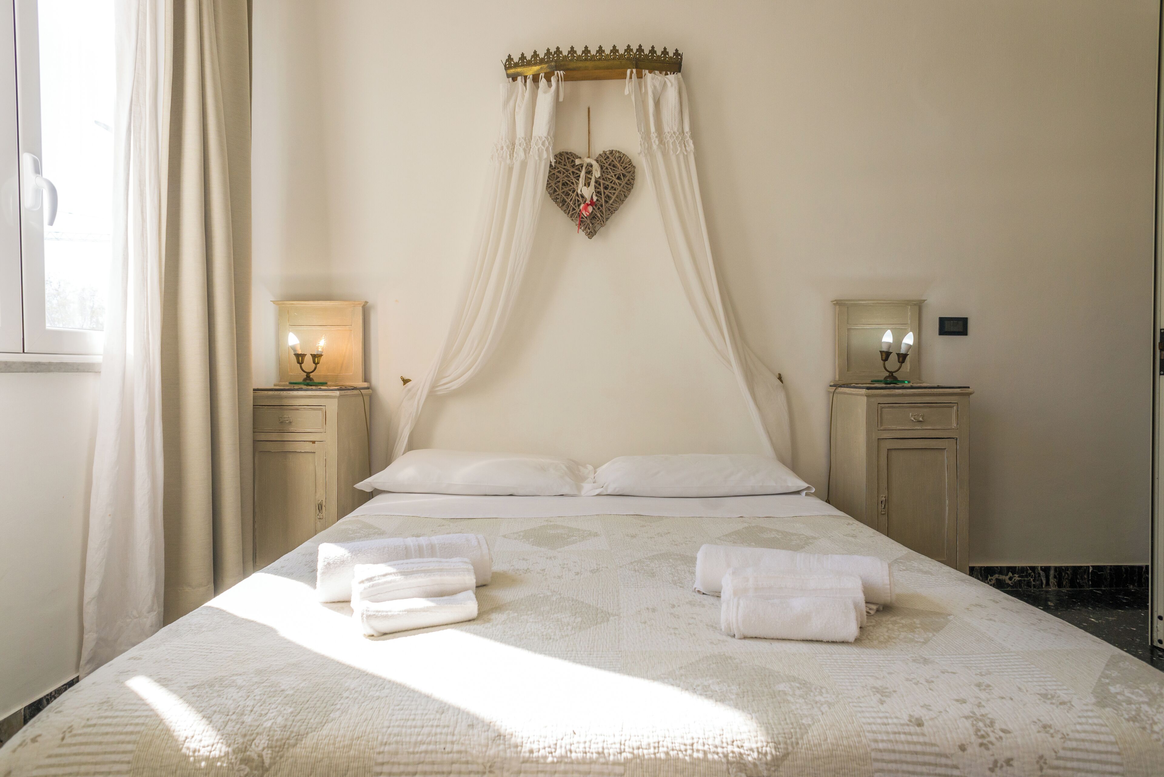 A neatly made bed with white linens and a heart-shaped decoration above it. There are two bedside tables with lamps and a small cabinet.