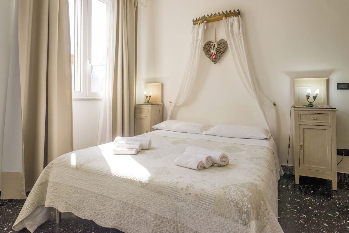 A neatly made bed with white linens and rolled towels, a bedside table with a lamp, and a decorative heart-shaped item on the wall.