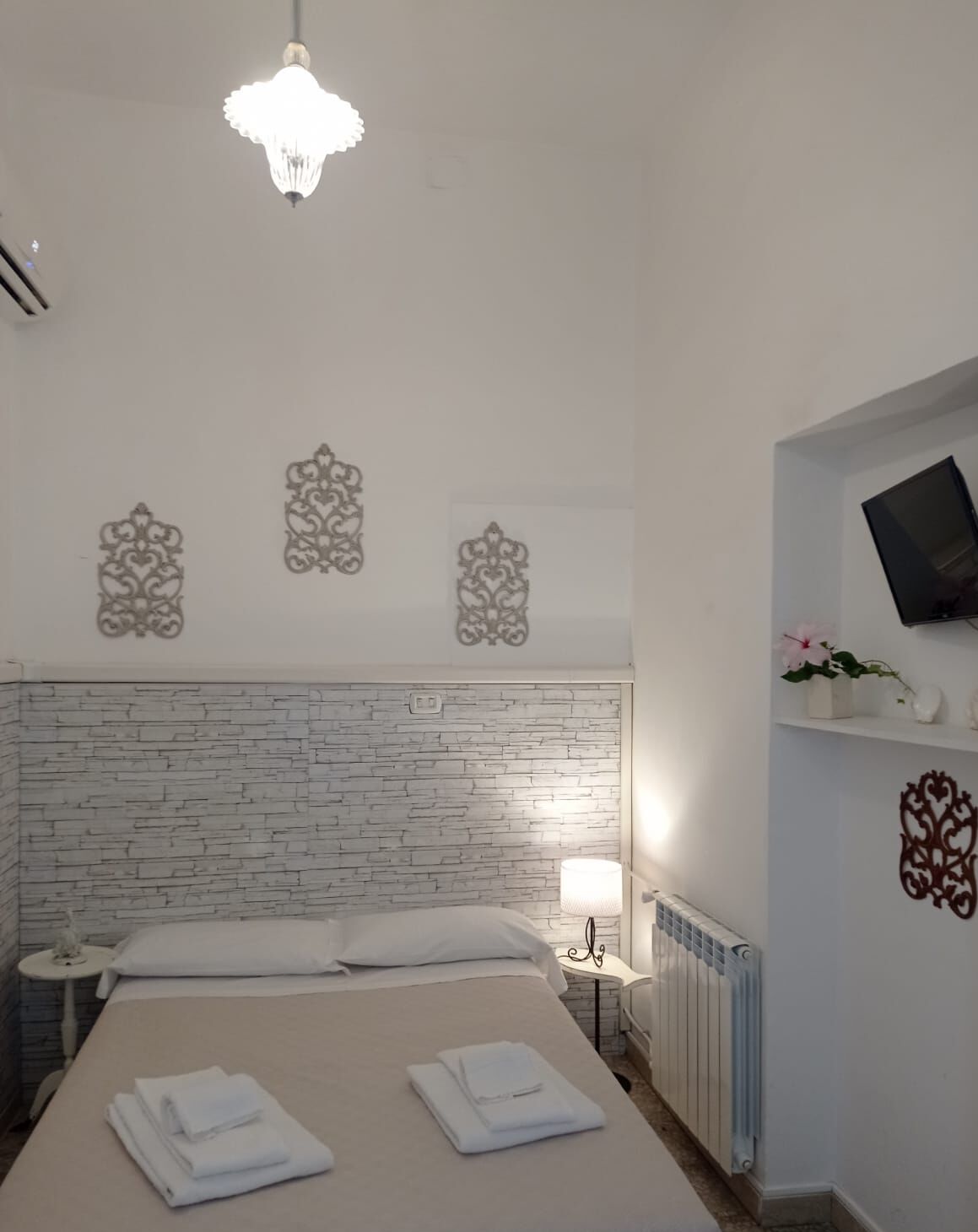 Ein Hotelzimmer mit Ziegelstein-Muster-Kopfstütze, einem Bett mit ordentlich gefalteten weißen Handtüchern, einem Nachttisch mit Lampe und einem an der Wand montierten Fernseher.