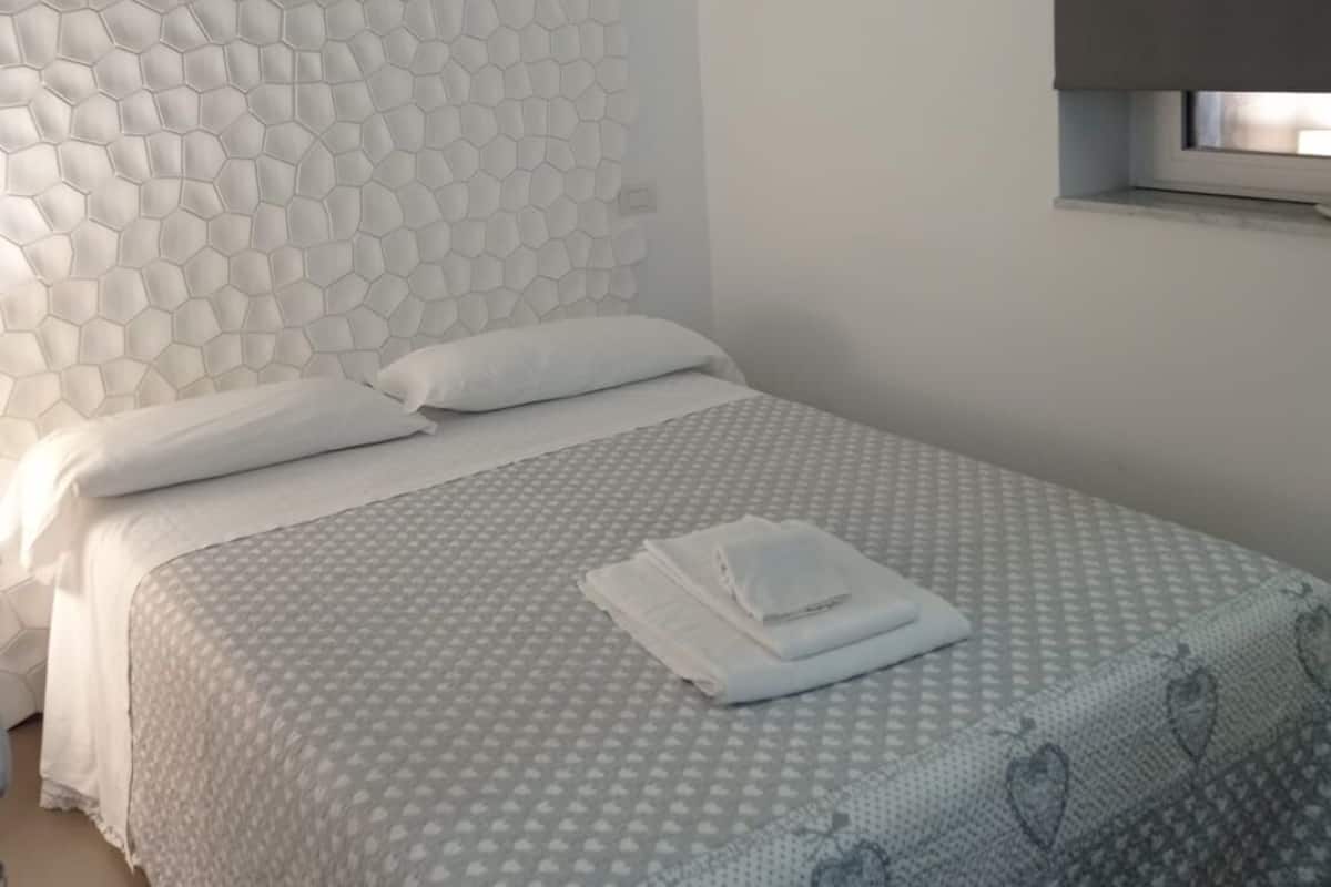 A neatly made bed with a patterned bedspread, a white headboard with a honeycomb design, and a window with a blind partially drawn.