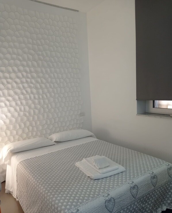 Een netjes opgemaakt bed met een bedsprei met patroon, een wit hoofdbord met een honingraatmotief, en een raam met een gedeeltelijk dichtgetrokken blind.