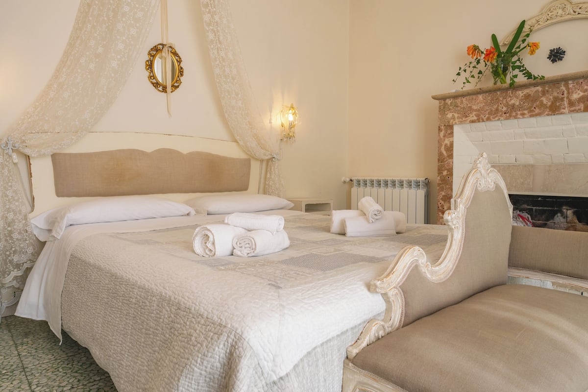 A bedroom with a large bed, a nightstand, a wall-mounted mirror, and a fireplace.