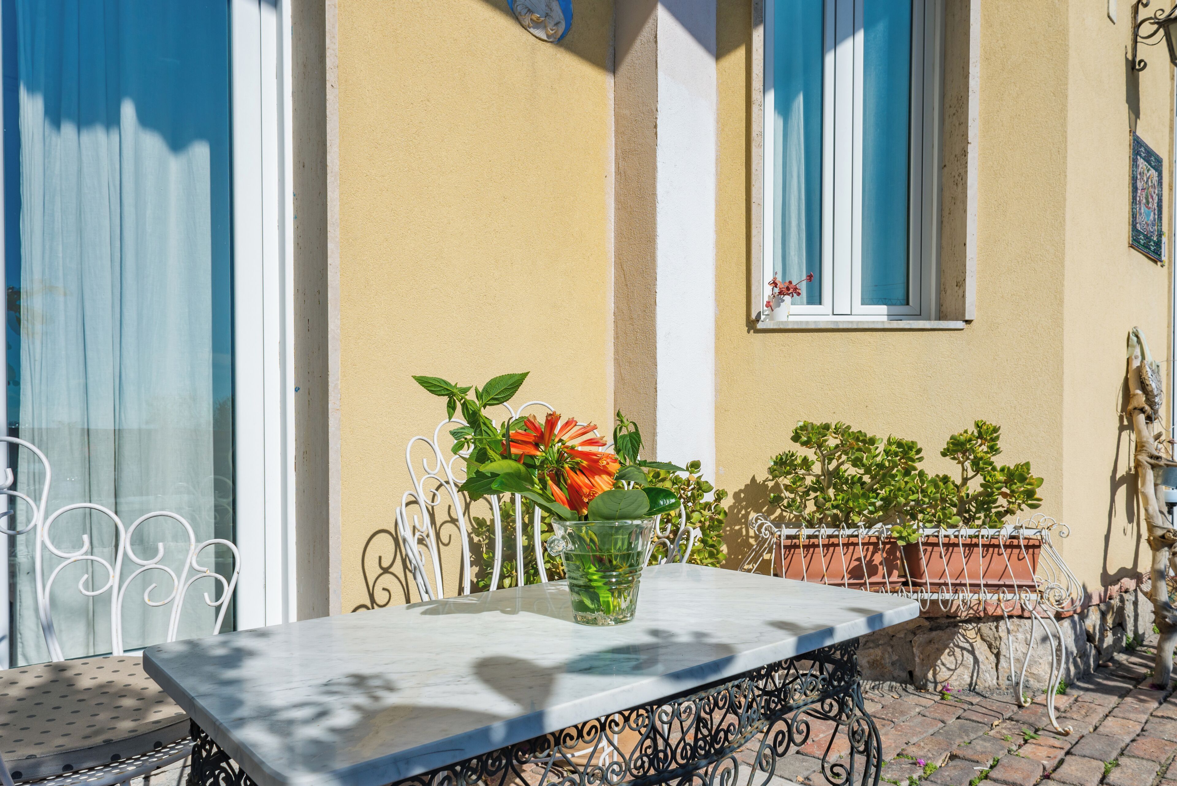 A table with a clear glass vase containing flowers, set on a balcony with potted plants and a window with blue shutters.