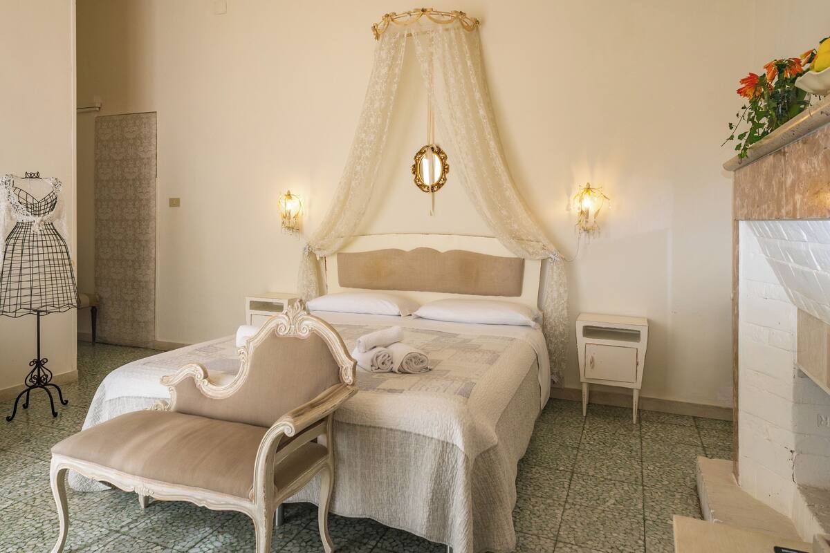 A bedroom with a canopy bed, two bedside tables, a chair, a lamp, and a wall-mounted mirror.