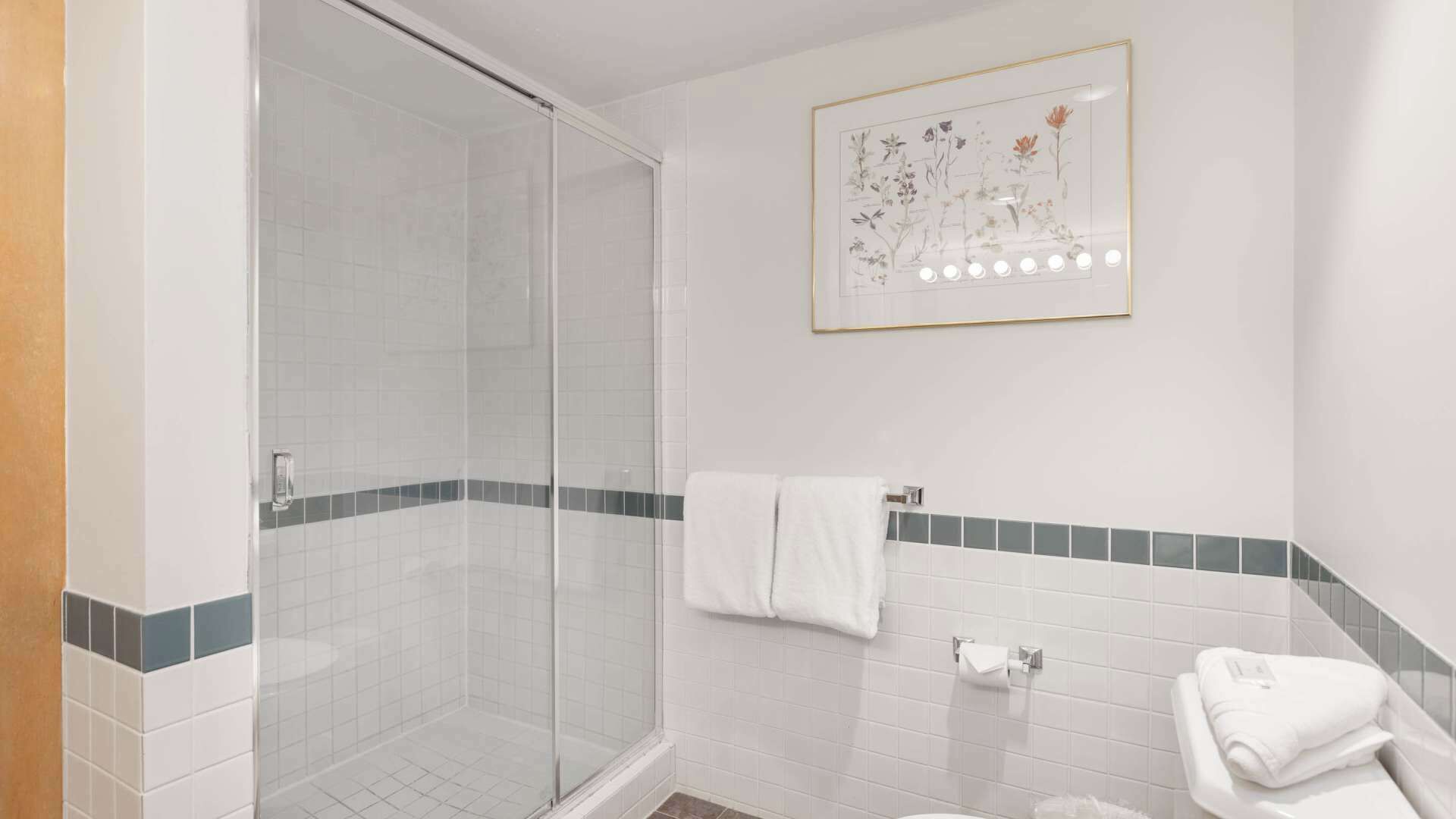 Un baño con cabina de ducha de vidrio, azulejos blancos, una ilustración botánica enmarcada y un perchero para toallas.