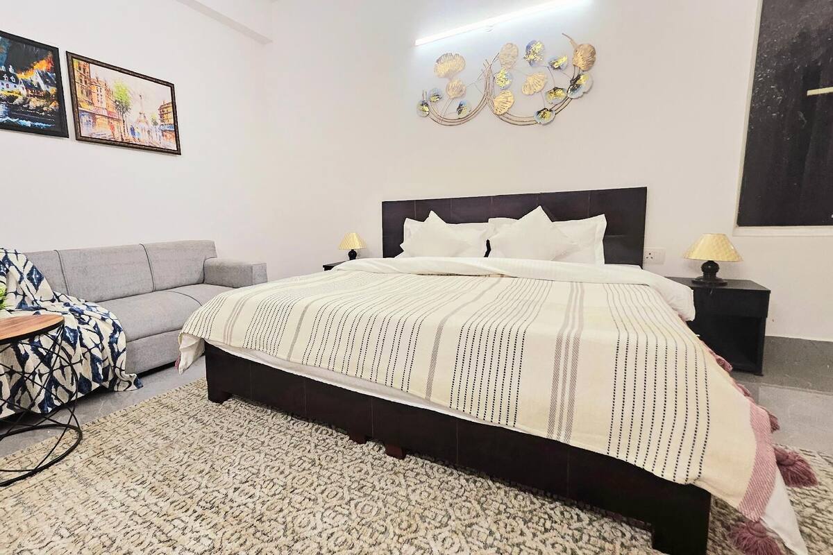 A bedroom with a bed, a grey sofa, a small table with a plant, and two wall art pieces.