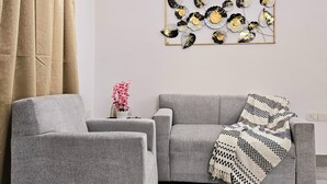 A living room with a grey sofa, two armchairs, a patterned throw blanket, a decorative wall piece, and a pink orchid vase. - Lux Studio Epic Stay (Gurugram)