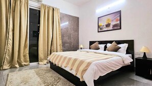 A bedroom with a bed, curtains, a painting, and a nightstand. - Lux Studio Epic Stay (Gurugram)