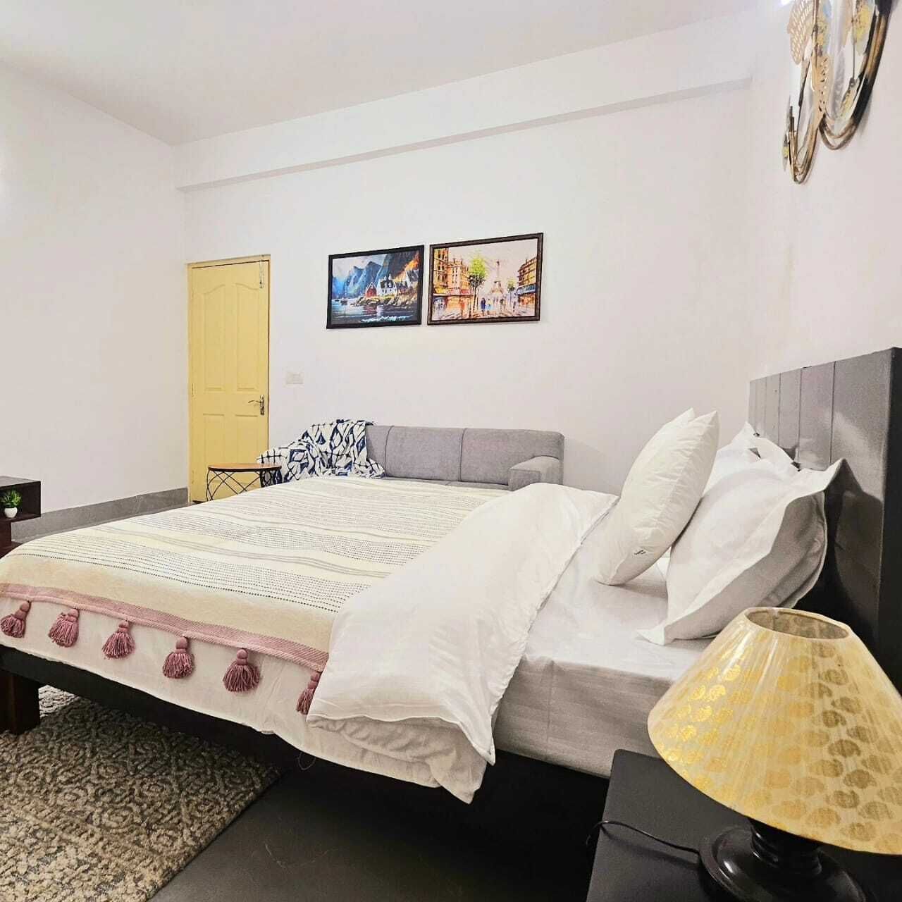 Un dormitorio con cama, lámpara de noche, alfombra y dos cuadros en la pared.