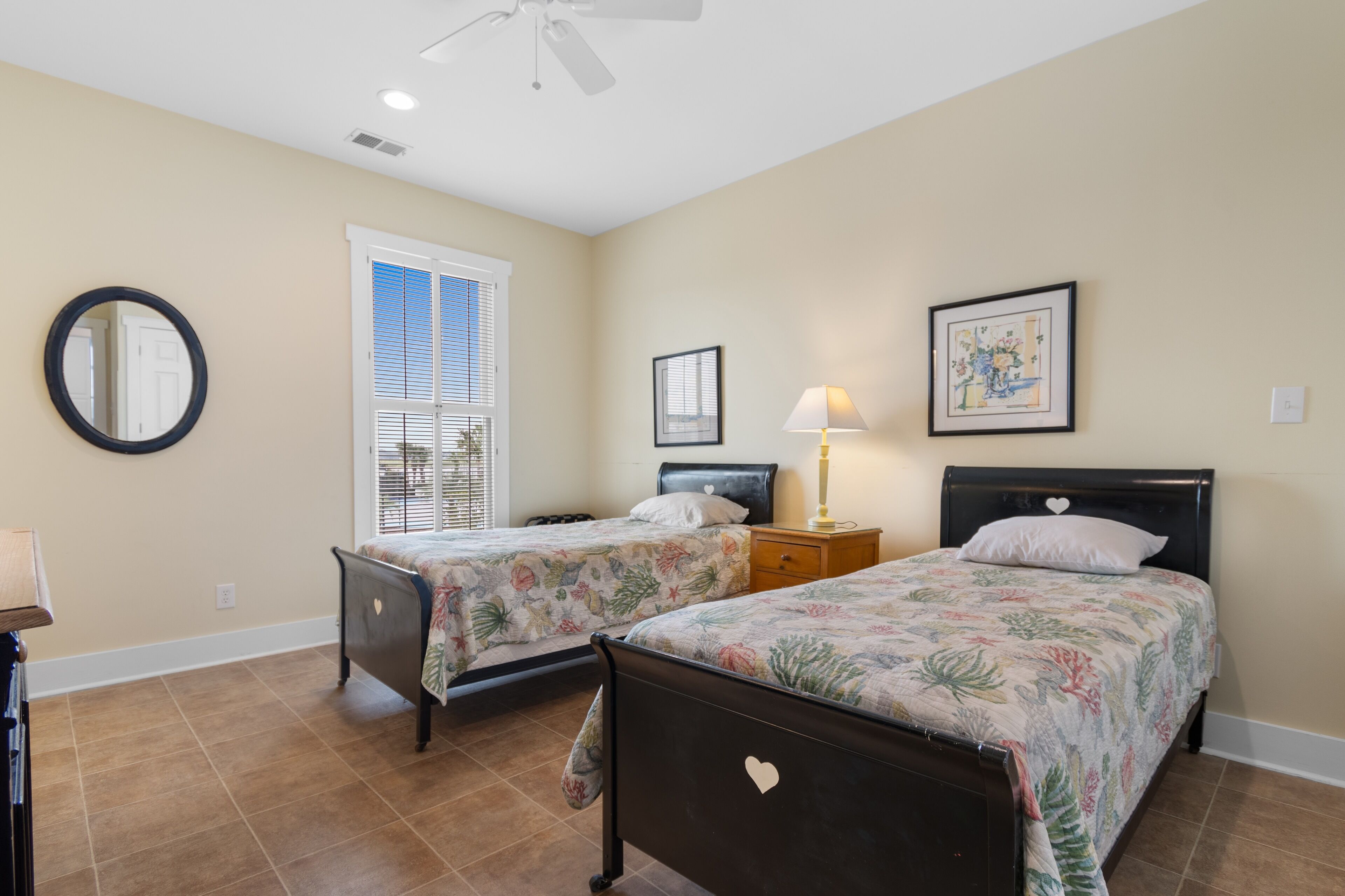 A bedroom with two beds, a ceiling fan, a window with blinds, and a framed picture on the wall.